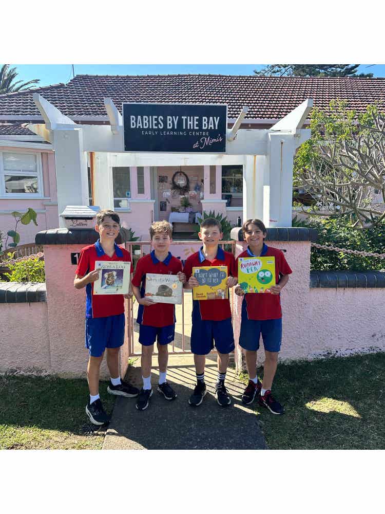 Four boys standing out the front of a 'Babies by the Bay' sign holding books and smiling at the camera.