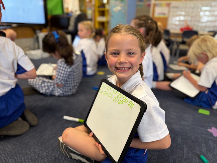 A girl sitting in on the floor in a classroom holding a whiteboard smiling at the camera.