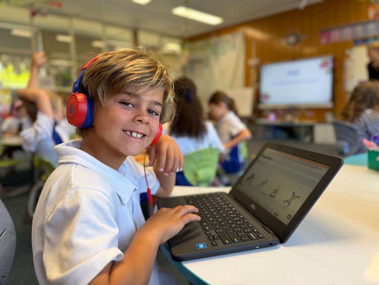 A student seated at a desk on the laptop with headphones on smiling at the camera.