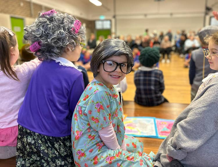 A student dressed as an old lady on the stage at an assembly with parent guests seated in the distant background.