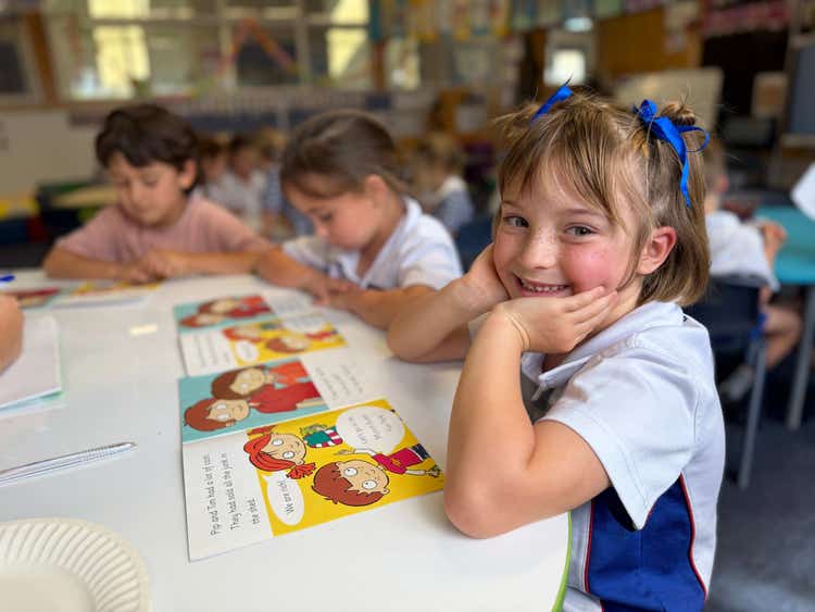 A student at a desk reading next to two other students with the student smiling at the camera.