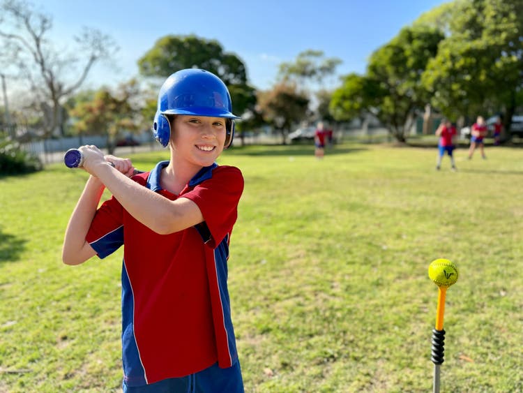 A boy holding a T-ball bat smiling at the camera ready to hit the ball off the tee with a green background.