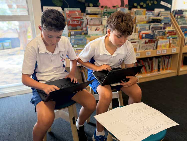 Two boys on laptop devices seated in a library with books in the background..