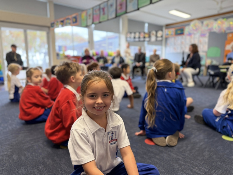 A kindergarten classroom with students seats on the floor, with the focus of one student facing the camera smiling.