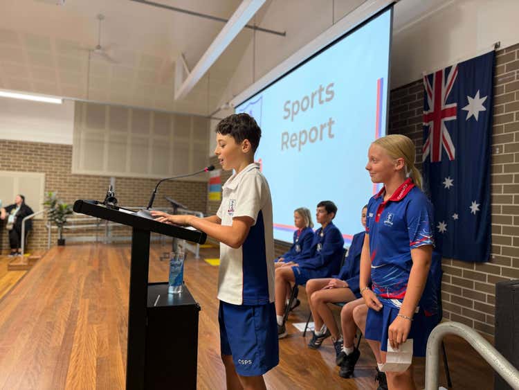 Student standing at a microphone on stage in a school hall addressing and audience.