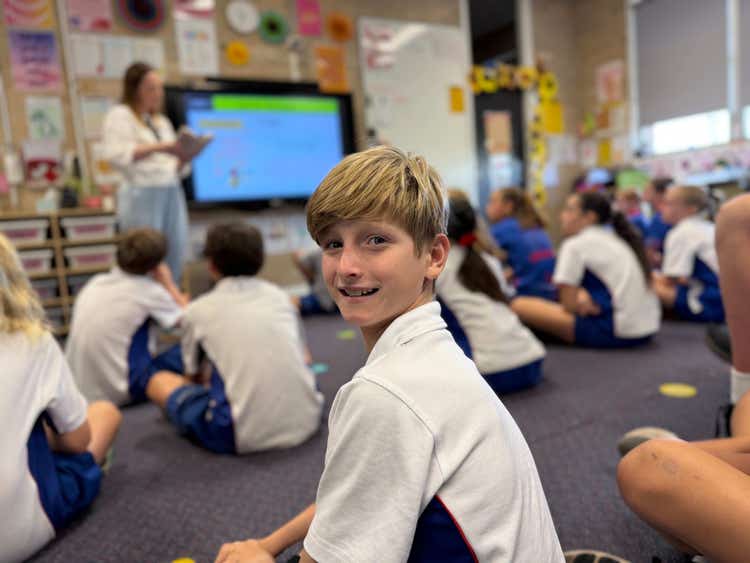 A stage 3 classroom with the teacher teaching and a student smiling at the camera.